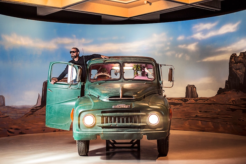 Colour photograph of artist Mark Shorter standing in a green truck in a gallery in front of a painted backdrop.