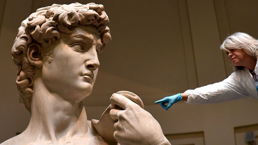 The head and shoulders of the statue of David, with a restorer, wearing a white coat, leaning in with their gloved hand.