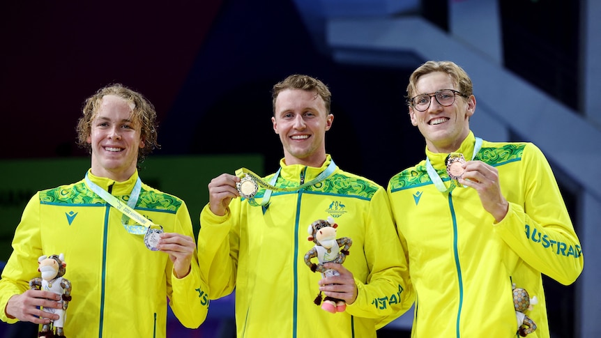 australian swimmers stand together on a podium holding medals all wearing matching yellow jackets