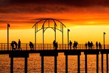 A pier above water with people walking on it at dusk is silhouetted against the bright orange glow of the background.