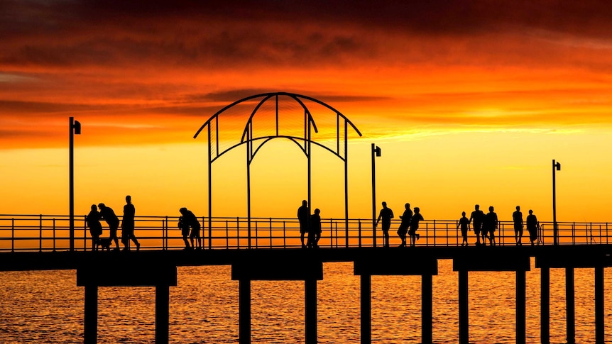 A pier above water with people walking on it at dusk is silhouetted against the bright orange glow of the background.