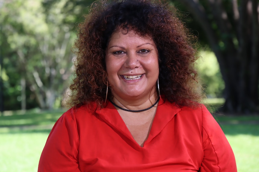 A woman wearing a red shirt smiles at the camera. She is outside in a park.