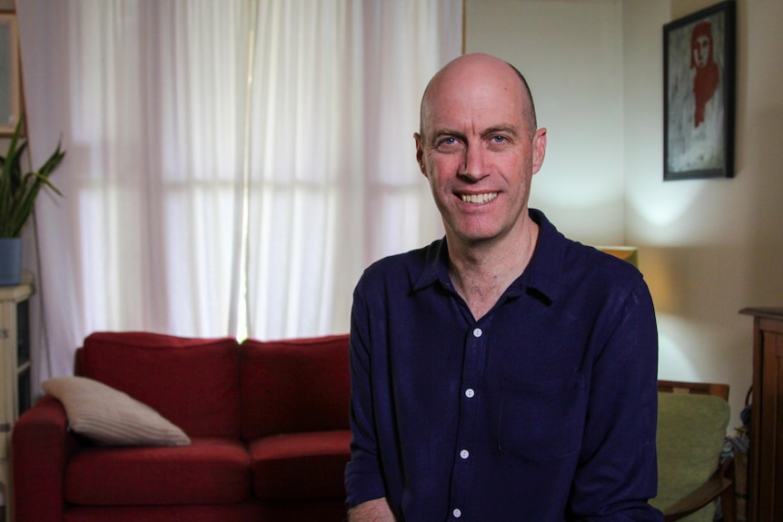 A middle age man sitting in a lounge room smiling at the camera