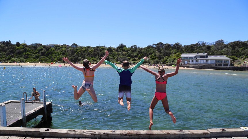 Three kids jump off a pier into water on a sunny day.