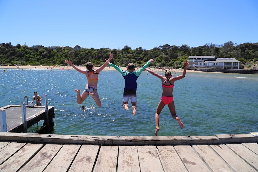 Three kids jump off a pier into water on a sunny day.