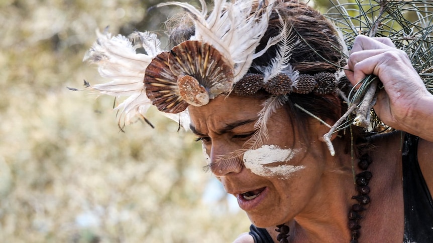 Close up of woman in traditional Indigenous headdress and face painted in ochre, dancing