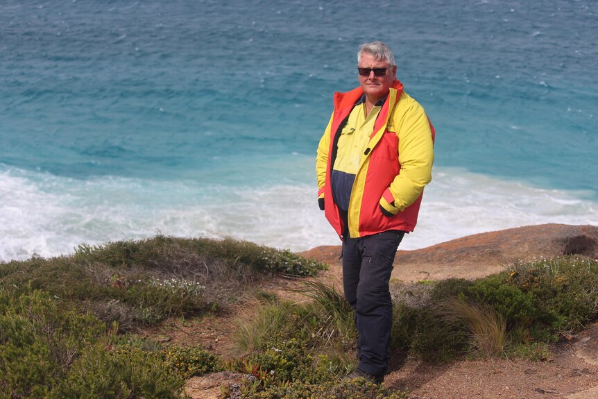 A full shot of a heavy set man in a yellow and orange jacket, standing on grassy land near blue water, white surf.