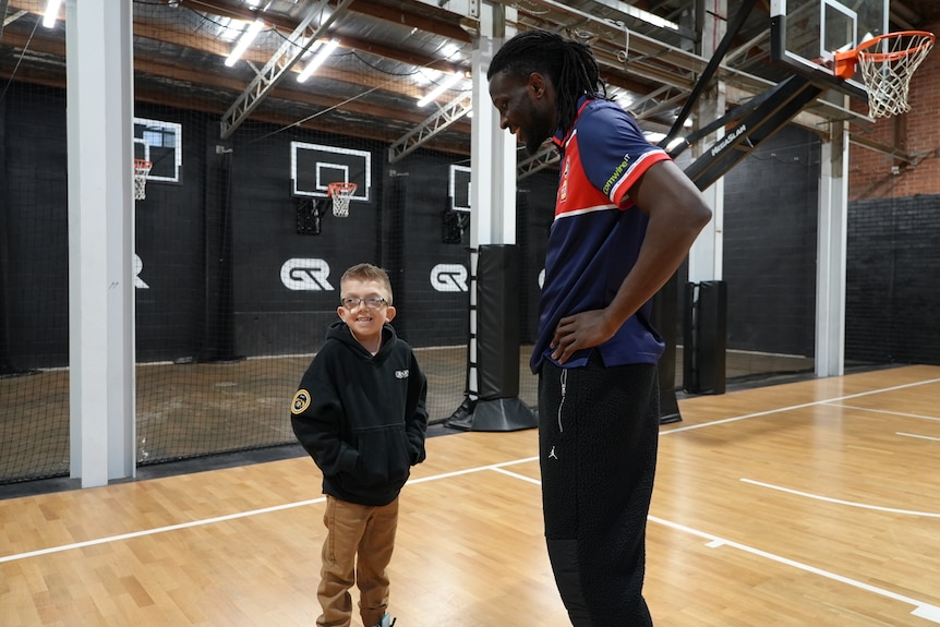 A tall basketball player stands next to a small young boy, they are both smiling