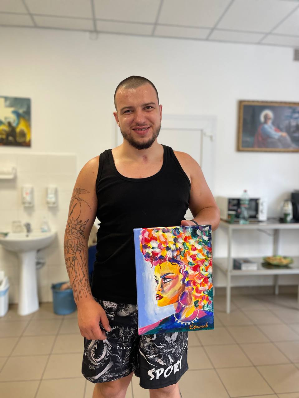A man with short hair and a beard wearing a black singlet holding up a colorful painting of a woman. 