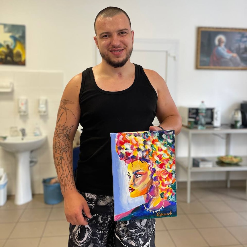 A man with short hair and a beard wearing a black singlet holding up a colorful painting of a woman. 