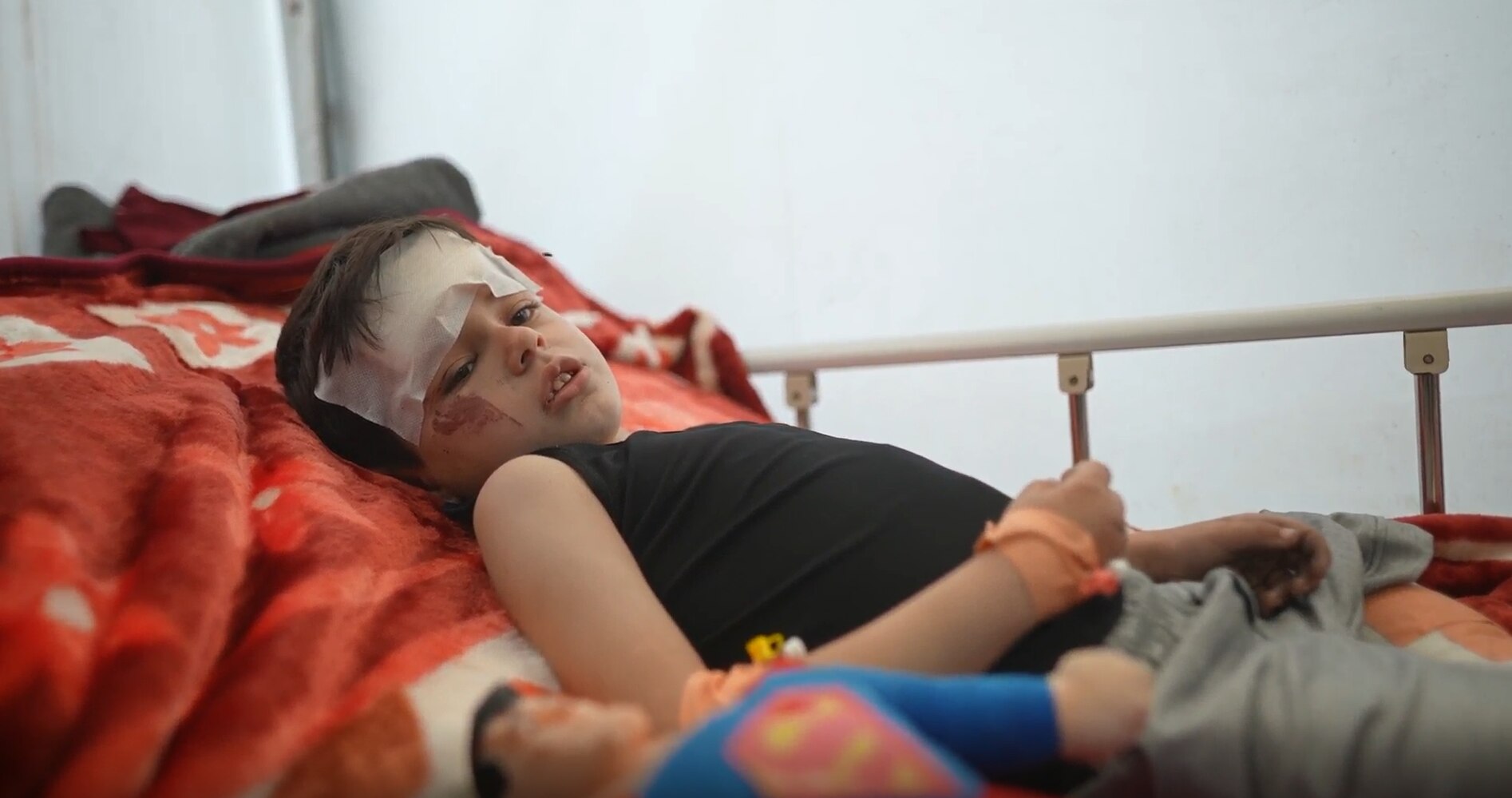 A boys sits on red blanket with a white bandage over his head 