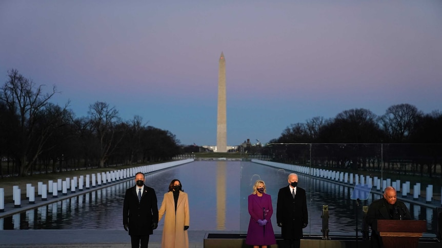 A man speaks at a lectern as two couples, all wearing masks, stand in front of the Reflecting Pool and the Washington Monument