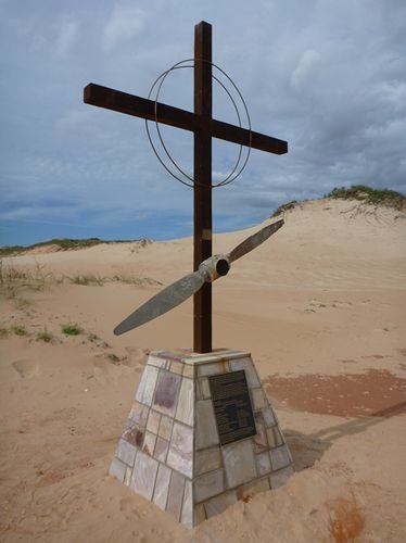 A large Christian cross mounted with a propeller stands on a monument in the sand.