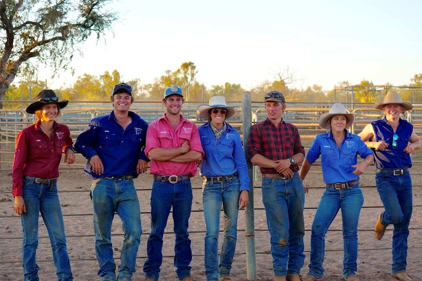 Seven stationhands wearing hats leaning against cattle yard fence