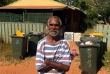 Indigenous man Rodney Wiggan stands outside a rundown house in Broome with lots of rubbish bins.