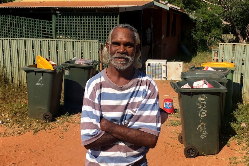 Indigenous man Rodney Wiggan stands outside a rundown house in Broome with lots of rubbish bins.