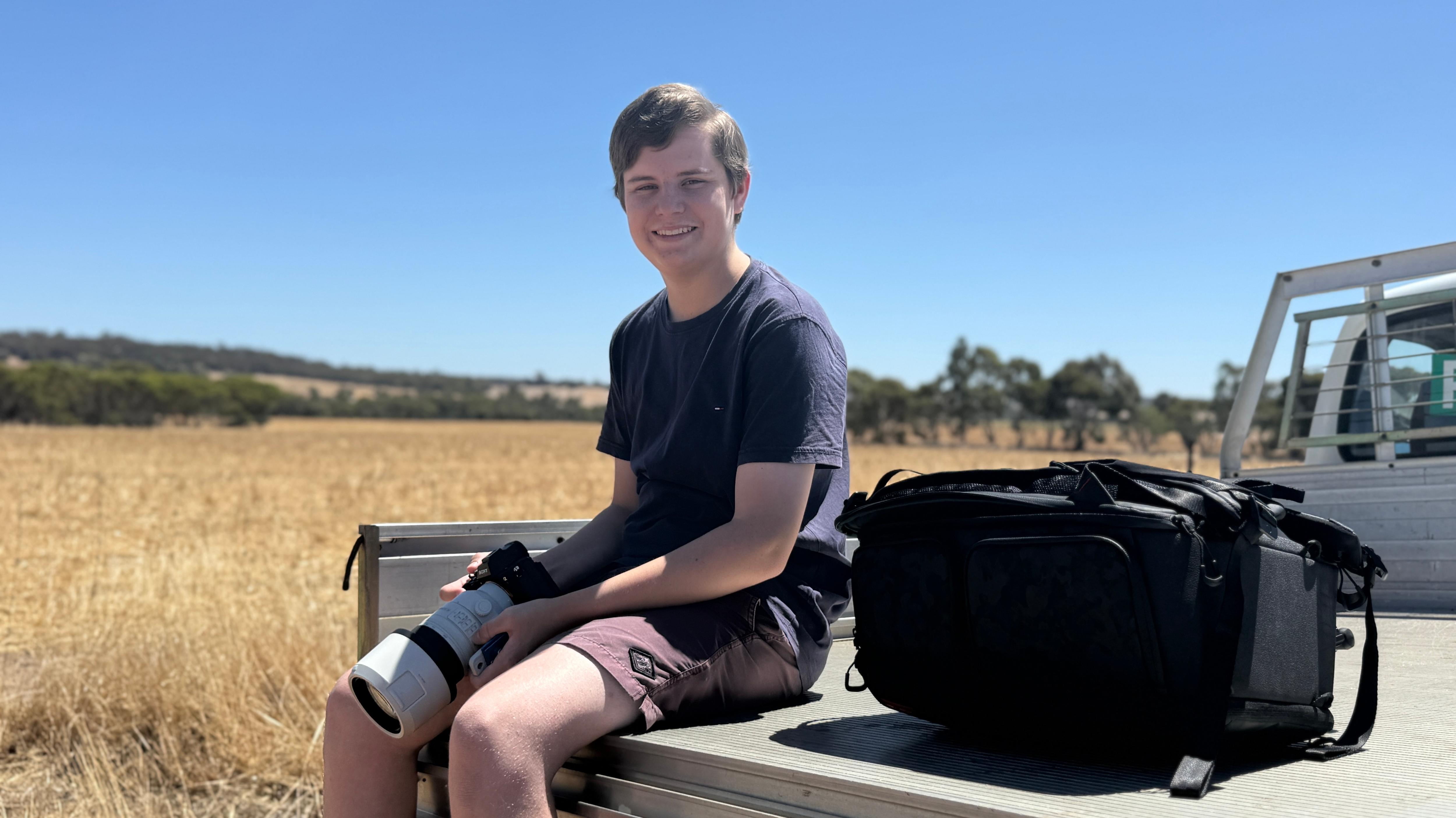 A smiling young man holds a camera with a telescopic lens as he sits on the back of a ute on a country property.