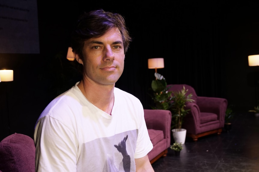 Matthew sits on a couch on stage, wearing a white t-shirt.