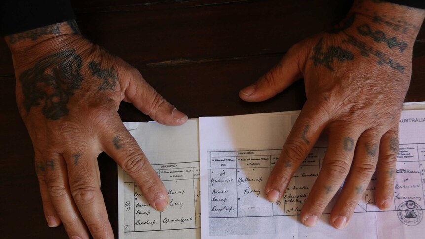 Tattooed hands show the difference between the original birth certificate with the label Aboriginal and the redacted version.