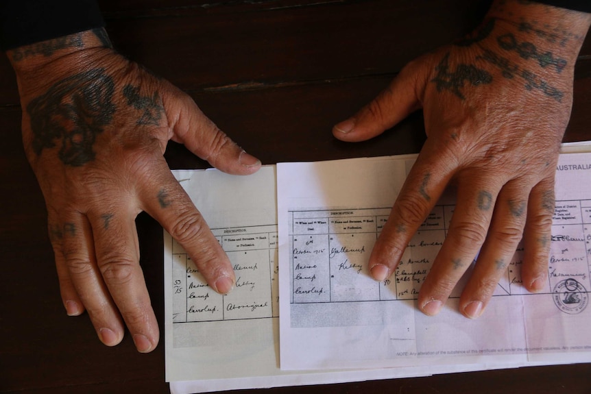 Tattooed hands show the difference between the original birth certificate with the label Aboriginal and the redacted version.