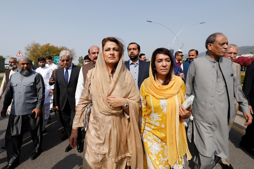 Two south Asian women in traditional flowing garb walk along city street with men behind them on sunny day