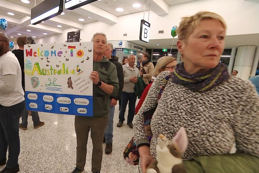 a man holding a sign that reads welcome to australia standing at the arrivals section of the airport