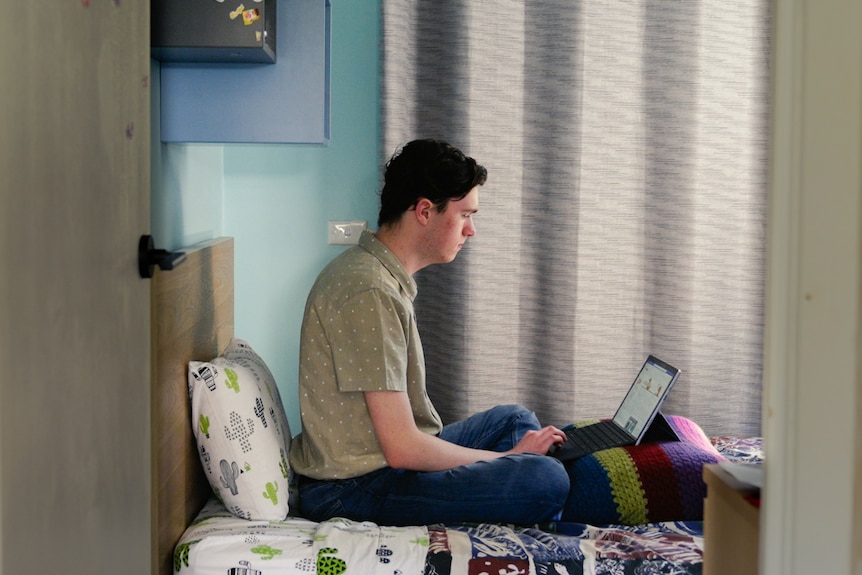 Jorden sits on his bed as he researches on his laptop. He wears a grey shirt and blue jeans and concentrates intently.