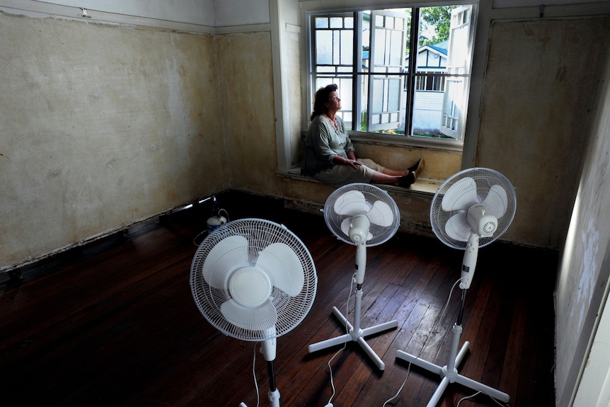 A pensive woman sits on her window seat and looks out.  Room is bare, walls are stained, has three pedestal fans.