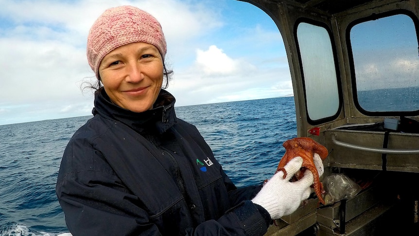 A woman wearing a pink beanie holds a red octopus on a boat