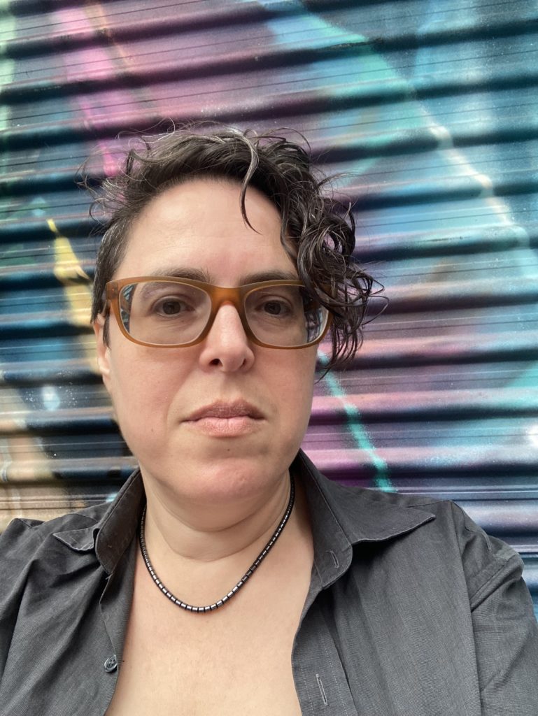 A person with a short asymmetrical curly hair cut and glasses stands in front of a bright graffitied roller door