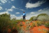 A traditional owner standing on top of a red sand dune with green flowers and shrubs lightly surrounding his feet