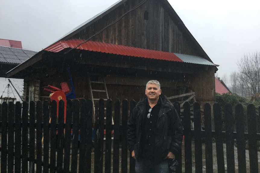 A man stands in front of a dark wooden cabin with red trims.
