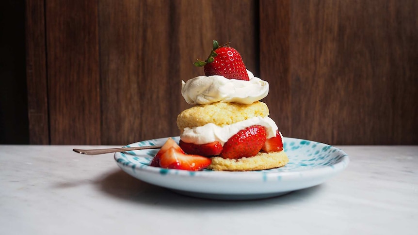 A strawberry shortcake sits on a plate topped with cream and a whole strawberry, an easy afternoon tea.