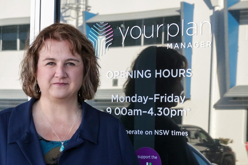 Serious looking woman with short hair looking into camera, standing outside a reflective window, with Your Plan written.