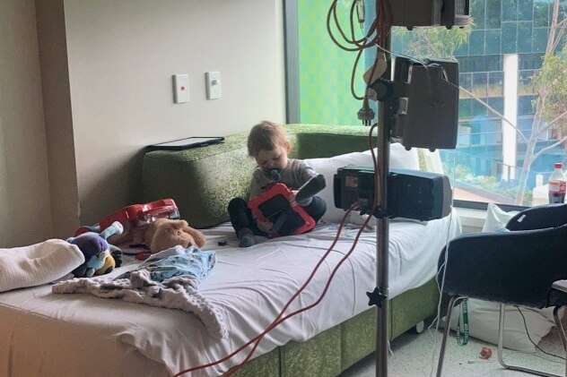 A little boy sits on a hospital bed with toys, hooked up to medical equipment. 