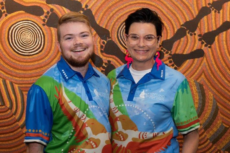 In front of an Indigenous painting of dots, circles and feet, a man with blonde hair and woman with brown hair stand smiling.