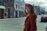 A young woman with curly red hair wears red beret and orange coat and stands in street looking up as snow falls on sunny day.