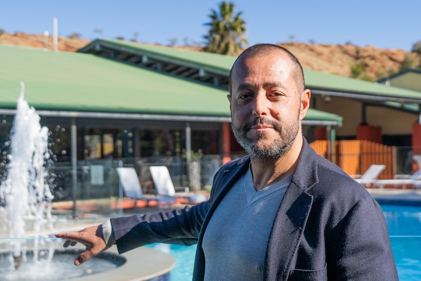 A smiling man with shaved hair, beard, stands by a pool, deck chairs, fountain behind. wears light blue tee and blue jacket.