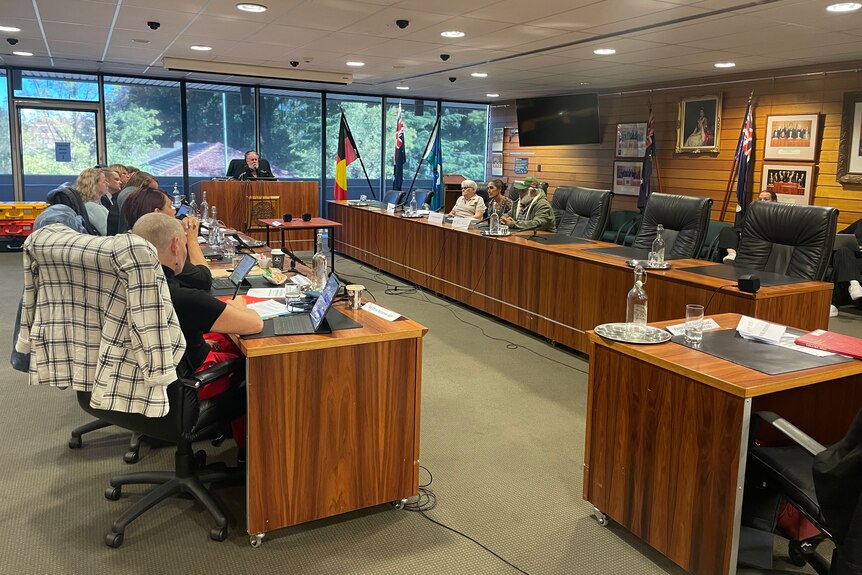 A group of people sit an a conference room, arrayed to face a chairman sitting next to some flags.
