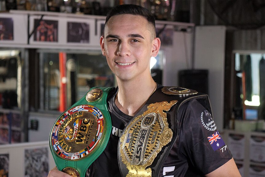 A young man with two boxing belts over his shoulders smiles at the camera.
