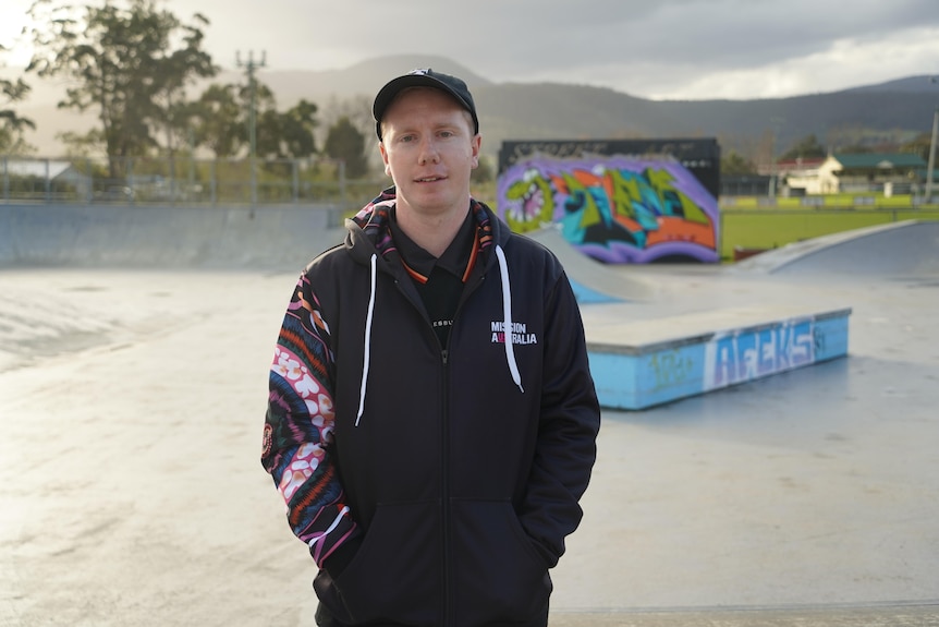 A young man in a baseball cap and hoodie stands in a skate park