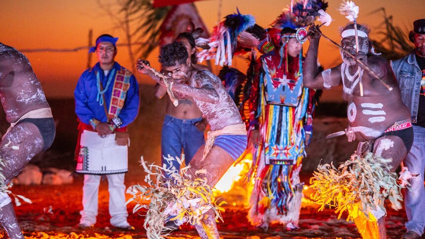 Indigenous dancers stand, snapped in action, dancing on the salt flats with the sunset behind them.