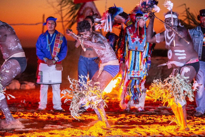 Indigenous dancers stand, snapped in action, dancing on the salt flats with the sunset behind them.