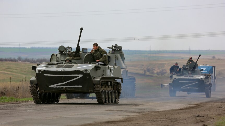 Three Russian military tanks with large letter Z on them, driving down a road amongst grass fields.