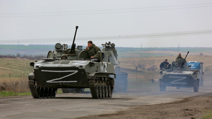 Three Russian military tanks with large letter Z on them, driving down a road amongst grass fields.