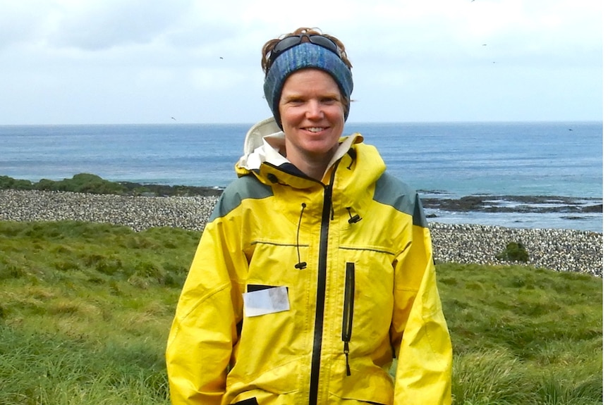 Woman in blue headscarf and yellow coat in front of beach with thousands of tiny white dots that are penguins