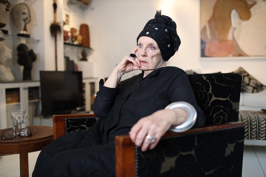 An elderly white woman dressed in black with a black headscarf sits in a dark armchair in a well appointed loungeroom.