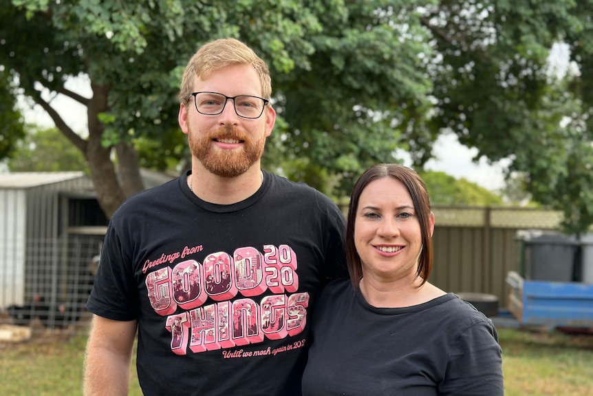 A smiling man and woman in t-shirts stand in a backyard with a large tree behind them.