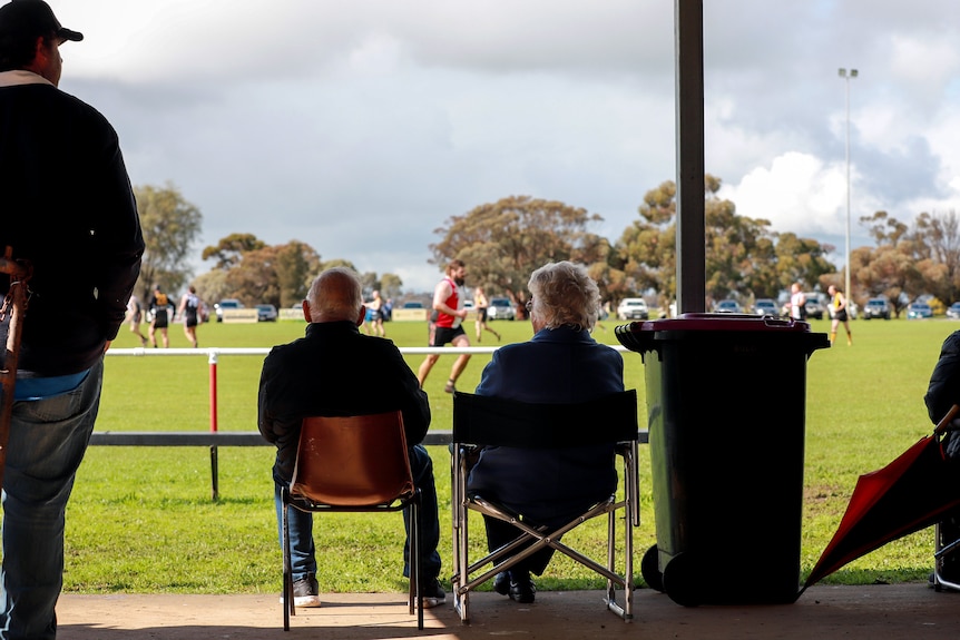 An older man and woman sit in chairs looking out toward a football oval with players running about