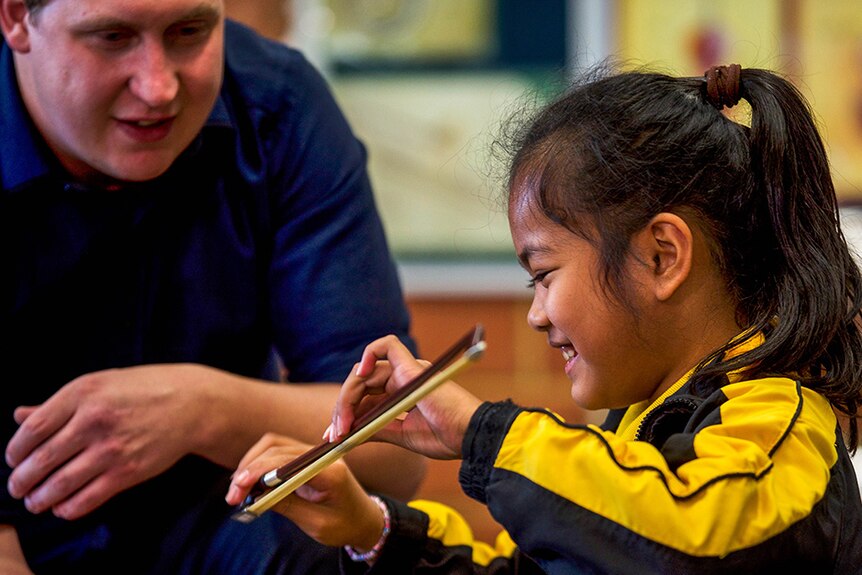 A schoolgirl holds a violin bow while a teacher in a blue shirt looks on